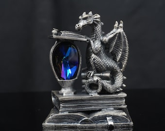 Puede incluir: Una figura de dragón plateado posada sobre una pila de libros, con un gran cristal azul profundo facetado incrustado en un pedestal. El dragón tiene escamas y alas texturizadas, y la base está formada por un libro abierto con detalles.