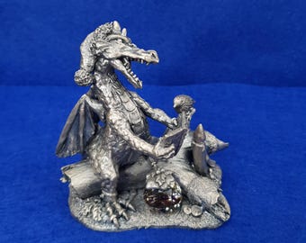 Puede incluir: Una figura de dragón de metal plateado con un gorro de Papá Noel, posada sobre un tronco. El dragón sostiene un libro abierto en sus garras, con una pequeña vela y un adorno decorativo con hojas de acebo descansando sobre el tronco.