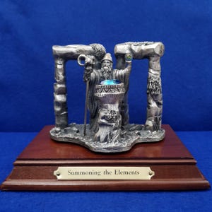 May include: A silver-toned metal figurine of a bearded wizard standing between two stone pillars, holding a staff with a ram head and a glowing orb. A blue crystal sits in a central basin. The base is wood with a plaque reading Summoning the Elements.
