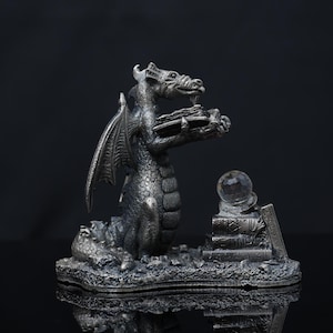 Op de afbeelding: Een zilverkleurig metalen drakenbeeldje op een getextureerde basis. De draak houdt een open boek in zijn klauwen, terwijl ernaast een stapel boeken staat met een heldere, bolvormige kristallen kogel erop. Het beeldhouwwerk heeft gedetailleerde schubben.