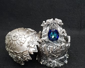 Puede incluir: Un joyero plateado de temática fantástica, con forma de huevo agrietado, con una gran gema azul. La caja presenta diseños de dragones y sol. La gema es de un azul intenso con toques de verde.