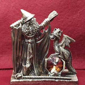 May include: A silver-colored figurine featuring a wizard with a long beard and a dragon. The wizard holds a book and stands next to the dragon, which is perched on a gem. The figurine is on a rectangular base.