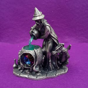 May include: A silver-toned metal figurine of a wizard wearing a pointed hat, pouring a translucent green liquid from a small pitcher into a cauldron. The cauldron features a large, multifaceted, iridescent blue and purple crystal gem.
