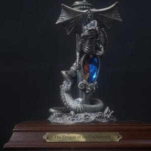 Puede incluir: Una figura de dragón plateada con alas extendidas enroscada alrededor de un pilar central con una piedra preciosa en forma de lágrima azul y naranja. La escultura se asienta sobre una base de madera con una placa de latón que dice The Dragon of the Underworld.