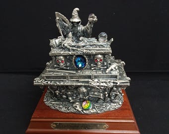 Puede incluir: Una escultura de metal plateado con una figura encapuchada que sostiene una guadaña y un pequeño orbe. La base es una estructura escalonada decorada con calaveras, huesos cruzados, una gran gema azul y una pequeña esfera iridiscente.