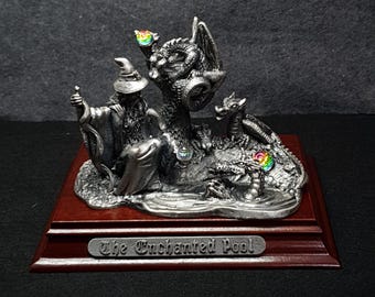 Puede incluir: Una detallada escultura de peltre que representa un mago, dragones y un pequeño dragón sobre una base. La escultura presenta detalles de cristal de colores y está montada sobre una base de madera oscura con el texto "The Enchanted Pool".