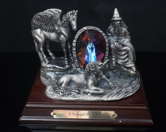 Puede incluir: Una figura de metal plateado sobre una base de madera oscura que muestra un caballo alado, un unicornio y una figura con túnica y sombrero puntiagudo. Un gran cristal ovalado facetado multicolor está en el centro. Placa de latón: A Tranquil Moment.