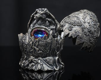 Puede incluir: Un recipiente metálico plateado en forma de huevo con tapa abatible. El exterior presenta relieves detallados de dragones y escenas forestales. Una gran piedra preciosa facetada de color púrpura y azul iridiscente está engastada en el centro.
