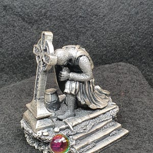 May include: A silver-colored figurine of a knight kneeling in prayer before a Celtic cross. The knight wears armor and kneels on a stone base. A colorful, iridescent gem is set at the base of the statue. A small barrel and sword are also present.