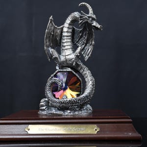 May include: A silver-toned dragon figurine perched atop a faceted, multi-colored crystal. The dragon is mounted on a rectangular dark wood base featuring a brass-colored plaque with the engraved text, The Guardian of the Crystal.