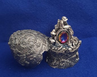 Puede incluir: Un joyero de metal plateado con tapa abatible. El exterior presenta intrincados relieves de dragones y follaje. En el interior, una figura esculpida se sitúa tras una piedra preciosa ovalada e iridiscente con tonos cambiantes de rojo, azul y dorado.