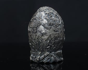 Puede incluir: Un objeto decorativo en forma de huevo de color plateado con intrincadas tallas en relieve de un dragón, un unicornio y un hada en un paisaje forestal. La superficie tiene un acabado metálico con texturas detalladas de alas, árboles y follaje.