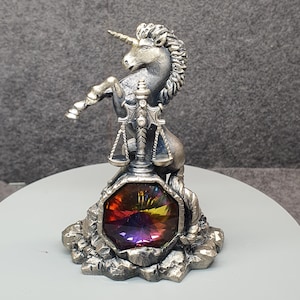 May include: A silver-colored unicorn figurine with a golden horn and a flowing mane. The unicorn is standing on its hind legs, with a colorful, faceted gemstone at its base. The gemstone displays a spectrum of colors, including red, yellow, and purple.