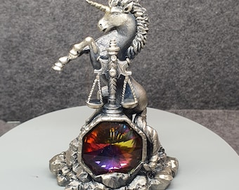 Puede incluir: Figura de unicornio plateado con cuerno dorado y melena detallada. El unicornio está sobre sus patas traseras, sobre una gema facetada multicolor. La gema muestra un espectro de colores, incluyendo rojo, amarillo y morado.
