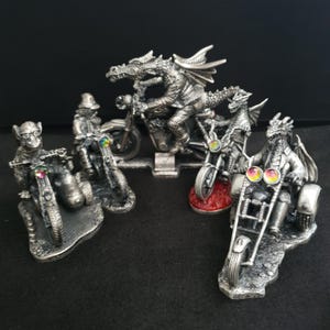 Puede incluir: Colección de figuras plateadas que representan varias criaturas, incluyendo dragones y un mono, montando motocicletas. Cada figura tiene detalles intrincados y está colocada sobre una superficie negra. Las motocicletas tienen faros coloridos con forma de joya.