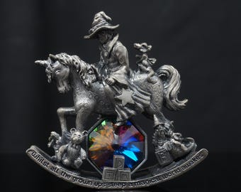 Puede incluir: Una figura de metal plateado de una persona con sombrero montando un unicornio, acompañada por una pequeña criatura. La base presenta un cristal facetado multicolor, un oso de peluche y el texto: whilst all the grown ups sleep a magical world awakes.