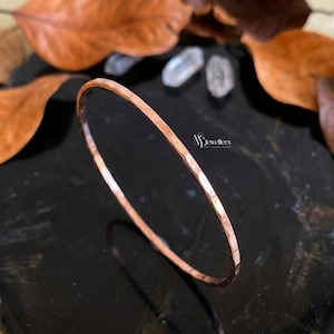 Könnte beinhalten: Ein gehämmertes Kupferarmband. Das Armband hat eine strukturierte Oberfläche und eine kreisförmige Form. Der Hintergrund umfasst braune Blätter und eine dunkle Oberfläche. Der Schmuck stammt von "VJ Jewellers".