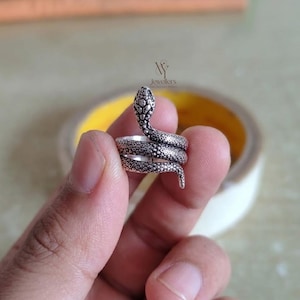Op de afbeelding: Een zilverkleurige slangenring met een gedetailleerd, getextureerd ontwerp. De kop van de slang is omhoog en het lichaam wikkelt zich om de vinger. De ring wordt in een hand gehouden, met het logo van de juwelier zichtbaar.