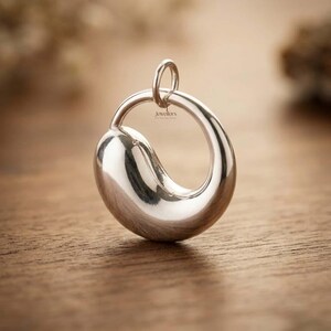May include: A silver pendant with a distinctive, flowing design. The pendant has a rounded, asymmetrical form with a loop at the top for a chain. The surface is highly polished, reflecting light. The pendant is presented on a wooden surface, with a blurred background.
