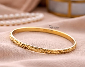 May include: A gold bangle with a detailed floral pattern. The bracelet is presented on a peach-coloured cloth. The jewellery is from WJ Jewellers.
