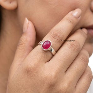 May include: A silver ring featuring a smooth, oval-shaped, deep pink cabochon gemstone set in a simple metal band. The text VYJewellers925 is visible on the right side of the ring. The band has a polished finish and a classic, clean design.