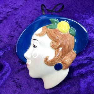May include: A ceramic wall hanging depicting a woman's profile wearing a blue hat. The woman has brown hair, a yellow flower, and a blue earring. The piece is set against a purple background and has a black ribbon for hanging.