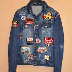 Może przedstawiać: Niebieska kurtka dżinsowa z wieloma naszywkami z tekstem i grafiką. Kurtka ma klasyczny kołnierz, zapięcia na guziki i białe plamy farby. Naszywki obejmują "DSQ Brothers" i "Mountain".
