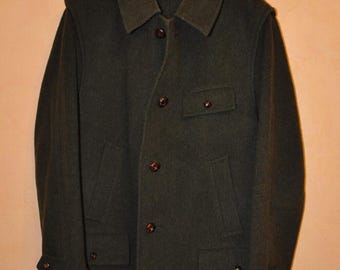 Può includere: Un cappotto di lana verde scuro dal design classico, con colletto a revers, chiusura con bottoni e più tasche. Il cappotto è appeso a una gruccia nera, mostrando la sua silhouette su misura e il tessuto resistente.