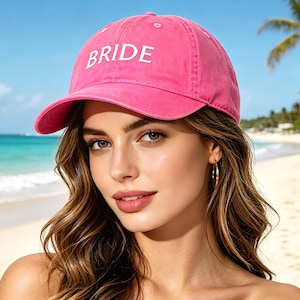 May include: A pink baseball cap with the word "BRIDE" embroidered in white. The cap is worn on a person's head, with a beach and palm trees in the background. The cap is made of a soft material and has a curved brim.