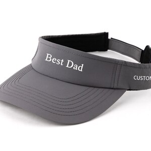 May include: Grey visor with the text "Best Dad" in white across the front. The word "CUSTOM" is printed on the side. The visor has a black interior and an adjustable strap.