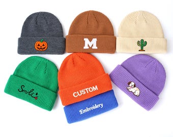 May include: A collection of six knitted beanies in various colors, including gray, brown, cream, green, orange, and purple. Each beanie features unique embroidered designs such as a pumpkin, the letter "M", a cactus, a dog, and the word "Smile". The orange beanie has the word "CUSTOM" and the blue beanie has the word "Embroidery".