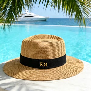 Peut inclure: Un chapeau fedora en paille beige doté d'un bord large et plat ainsi que d'une calotte structurée. Un ruban en gros-grain noir entoure la base de la calotte, orné des lettres capitales dorées KG centrées sur le devant du ruban.