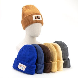 Peut inclure: Plusieurs bonnets en tricot de couleurs variées, dont marron, bleu et gris, exposés sur une tête de mannequin. Le bonnet marron porte un petit écusson avec une photo. Le bonnet bleu porte un écusson avec un mouton et le texte "happy sheep days".