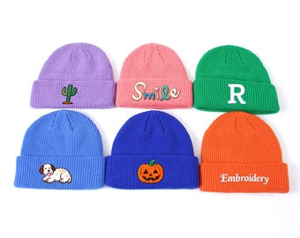 May include: Six colorful knit beanies are displayed. The beanies are in shades of purple, pink, green, blue, and orange. Each beanie has a different embroidered design, including a cactus, the word "Smile", the letter "R", a dog, a pumpkin, and the word "Embroidery".