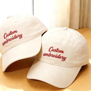 May include: Two cream-colored baseball caps with curved visors are displayed. Each cap has the words "Custom embroidery" embroidered in red script across the front. The caps are resting on a wooden surface.