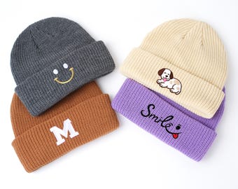 May include: Four knit beanies in various colors: gray with a yellow smiley face, brown with a white "M", cream with a puppy, and purple with the word "Smile" and a red tongue. These beanies are perfect for cold weather.
