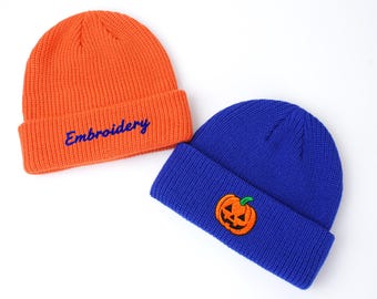 May include: Two knit beanies. One is orange with the word "Embroidery" in blue. The other is blue with an embroidered jack-o'-lantern patch. Both beanies have a folded brim and are made of a ribbed knit material.