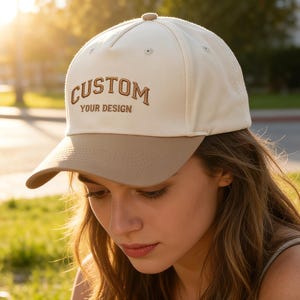 Puede incluir: Gorra de béisbol beige con visera marrón. La gorra tiene el texto bordado "CUSTOM YOUR DESIGN" en una fuente marrón estilizada. La gorra la lleva una persona al aire libre con luz natural.