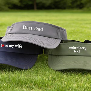 Könnte beinhalten: Drei Visiere mit Text auf einer grasbewachsenen Oberfläche. Das obere Visier ist grau und trägt die Aufschrift "Best Dad". Das mittlere Visier ist marineblau und trägt die Aufschrift "I love my wife". Das untere Visier ist olivgrün und trägt die Aufschrift "embroidery text".