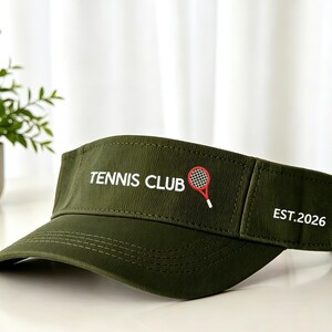 May include: An olive green visor featuring the white text TENNIS CLUB, a small red and white tennis racket graphic, and the white text EST.2026 on the side panel. The visor has a curved brim with visible stitching and an adjustable strap.
