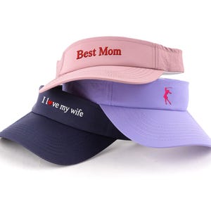 May include: Three visors in pink, lilac, and navy blue are stacked. The pink visor has "Best Mom" embroidered in red. The navy visor says "I love my wife" in white. The lilac visor has a red golf swing figure.