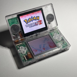 May include: A transparent Nintendo DS handheld game console, open to display the game "Pokémon White Version 2." The top screen shows the game's logo in white, blue, and red. The bottom screen displays in-game graphics. The console's buttons and internal components are visible.