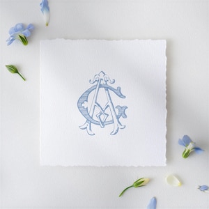 May include: A light blue monogram design featuring the letters "A" and "G" intertwined, printed on a square, textured white paper with a deckled edge. Blue flowers and buds are scattered around the paper.