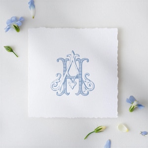 May include: A light blue, ornate monogram design featuring the letters "A" and "H" on a square, textured white paper. The paper has a deckled edge. Scattered around the paper are small blue flowers and buds, creating a delicate, floral arrangement.