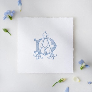 Puede incluir: Un diseño de monograma azul claro con las letras entrelazadas "DA" impresas en un papel blanco cuadrado texturizado con un borde dentado. Flores y capullos azules están dispersos alrededor del papel, creando una estética delicada y elegante.
