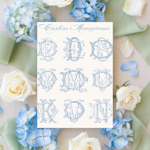 Op de afbeelding: Een crèmekleurige kaart met negen lichtblauwe aangepaste monogrammen, met de tekst "Custom Monograms" bovenaan. De kaart is omgeven door witte rozen, blauwe hortensia's en groene linten.