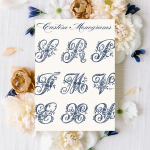 May include: A card featuring custom monograms in a navy blue script font. The monograms are ornate, with floral embellishments. The card is surrounded by white and cream-colored flowers and petals on a white background.