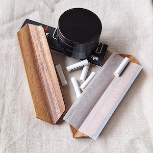 May include: A collection of smoking accessories, including a black grinder, wooden rolling tray, rolling papers, and white filter tips. The items are arranged on a neutral-colored surface, with the wooden tray and accessories providing a natural aesthetic.