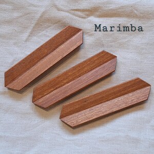 May include: Three wooden marimba bars with a two-tone design. The bars are a medium brown with a lighter brown center stripe. The word "Marimba" is printed in black above the bars. The bars are arranged diagonally on a light beige surface.