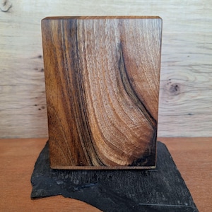 May include: A rectangular wooden box with a rich, dark brown grain pattern. The box is set on a dark, textured, black stone base. The wood grain has a natural, swirling pattern.